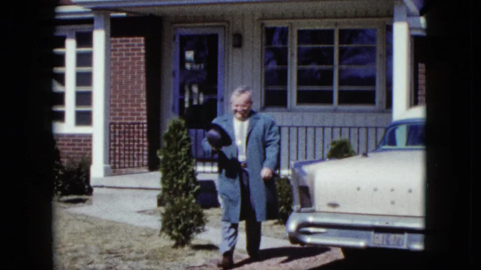 The man continues along the residential walk, carrying his hat as the front and side of a pale vintage car fill the right side of the frame.
