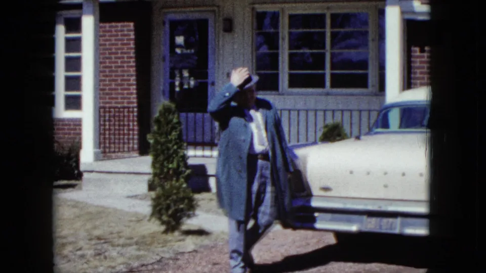 The man lifts his brimmed hat near the parked car, with the brick-and-siding Nevada house visible behind him in the 1959 home movie.