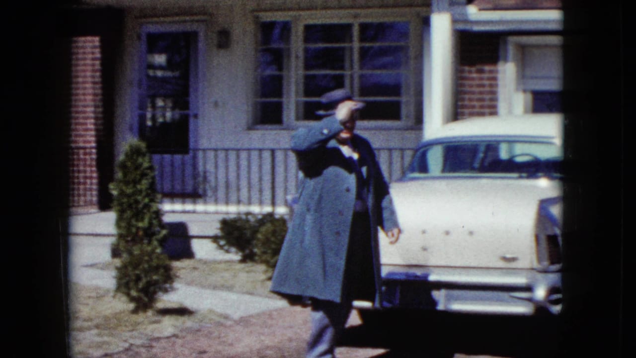 Man in a fedora and long coat waves beside a light-colored sedan in a driveway.