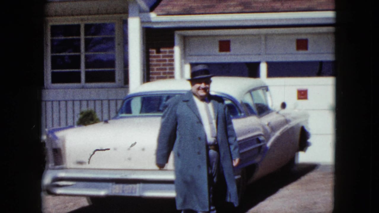 Smiling man in a dark hat stands near a light-colored sedan and garage door.