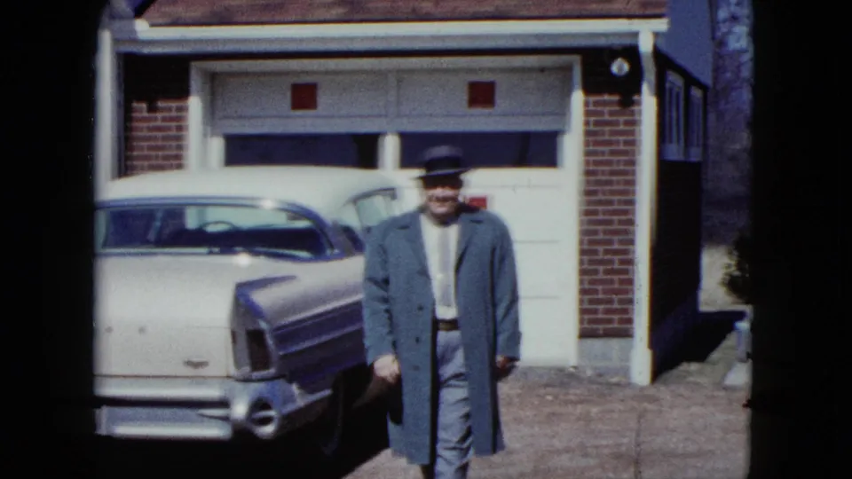 The man faces the camera in a driveway, with a mid-century sedan and garage door behind him in Nevada home-movie footage.