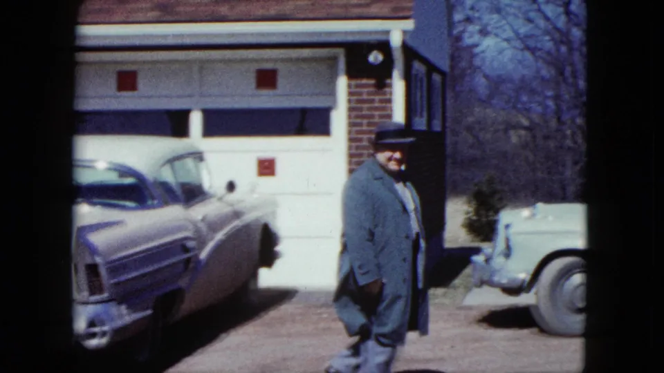 A man in a fedora and long coat walks near a driveway, with a 1950s sedan and another car visible in the frame.