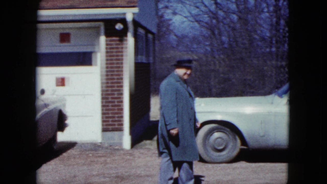 Man in a fedora walks past a garage toward a light-colored car in a driveway.