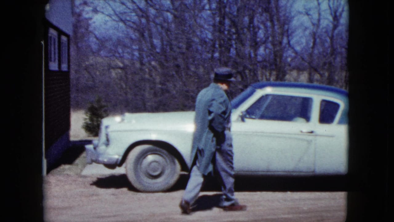 A man in a fedora and coat walks beside a light-colored car with a blue roof in a Nevada driveway, with bare trees behind him.