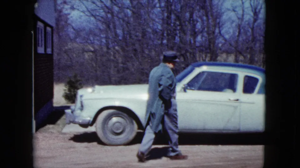A man in a fedora and coat walks beside a light-colored car with a blue roof in a Nevada driveway, with bare trees behind him.