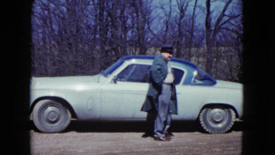 The man pauses next to the driver-side area of a light-colored mid-century car in 1959 Nevada home-movie footage.