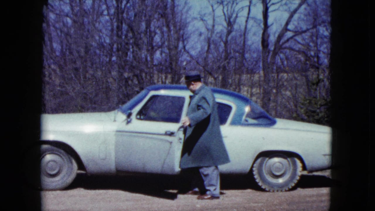 In 1959 Nevada home-movie footage, an older man in a coat and hat opens the driver's door of a light-colored mid-century car.