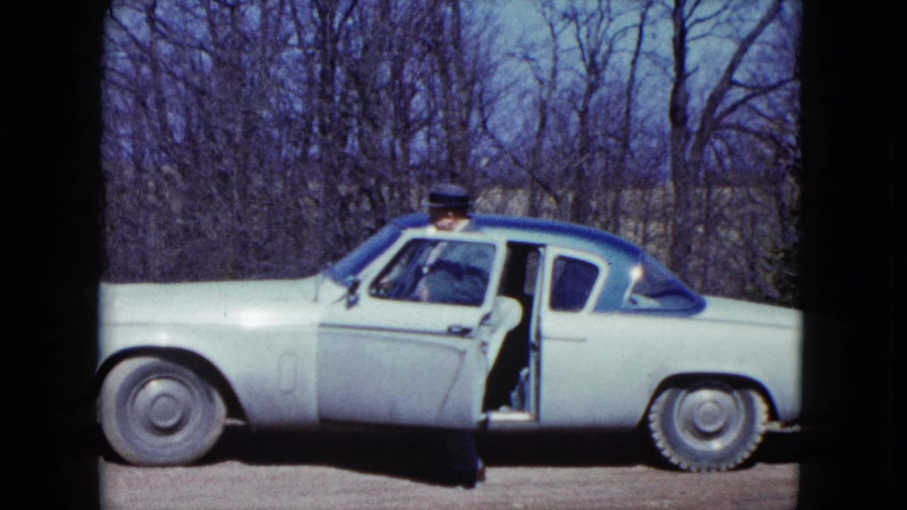 The older man steps into the open driver's side of the pale two-tone car, with leafless trees behind him.