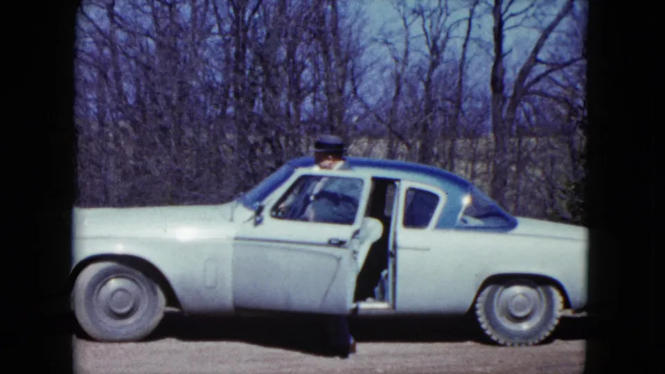 The older man steps into the open driver's side of the pale two-tone car, with leafless trees behind him.