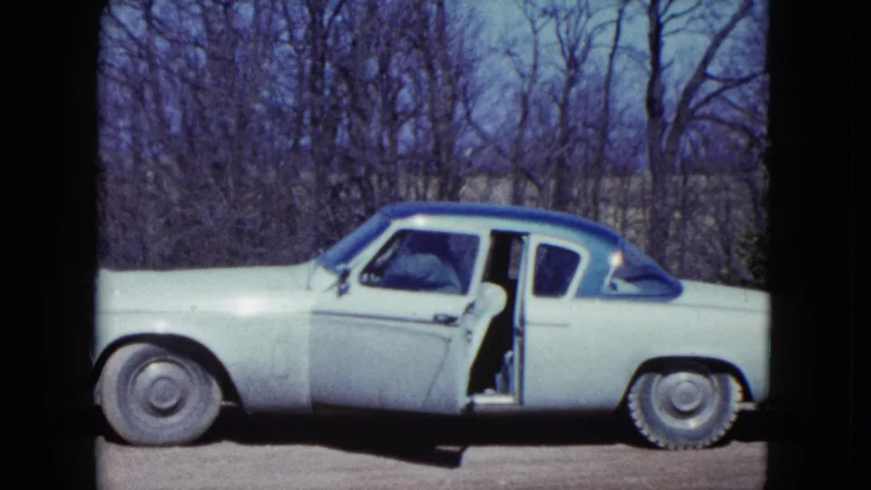 The car remains framed broadside in the wooded Nevada setting, its driver's door open as the man moves inside.