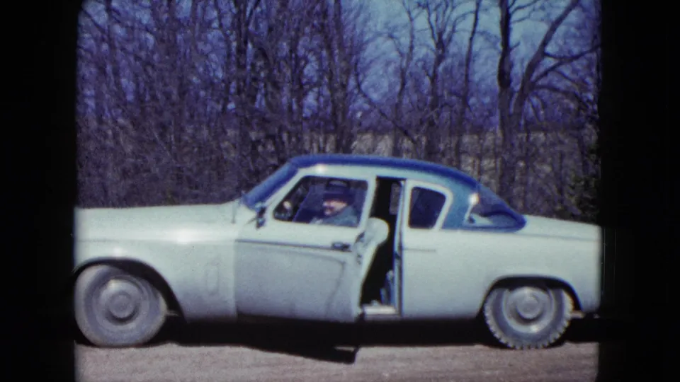 The older man sits in the driver's seat of the pale mid-century car, visible through the window with the door still ajar.