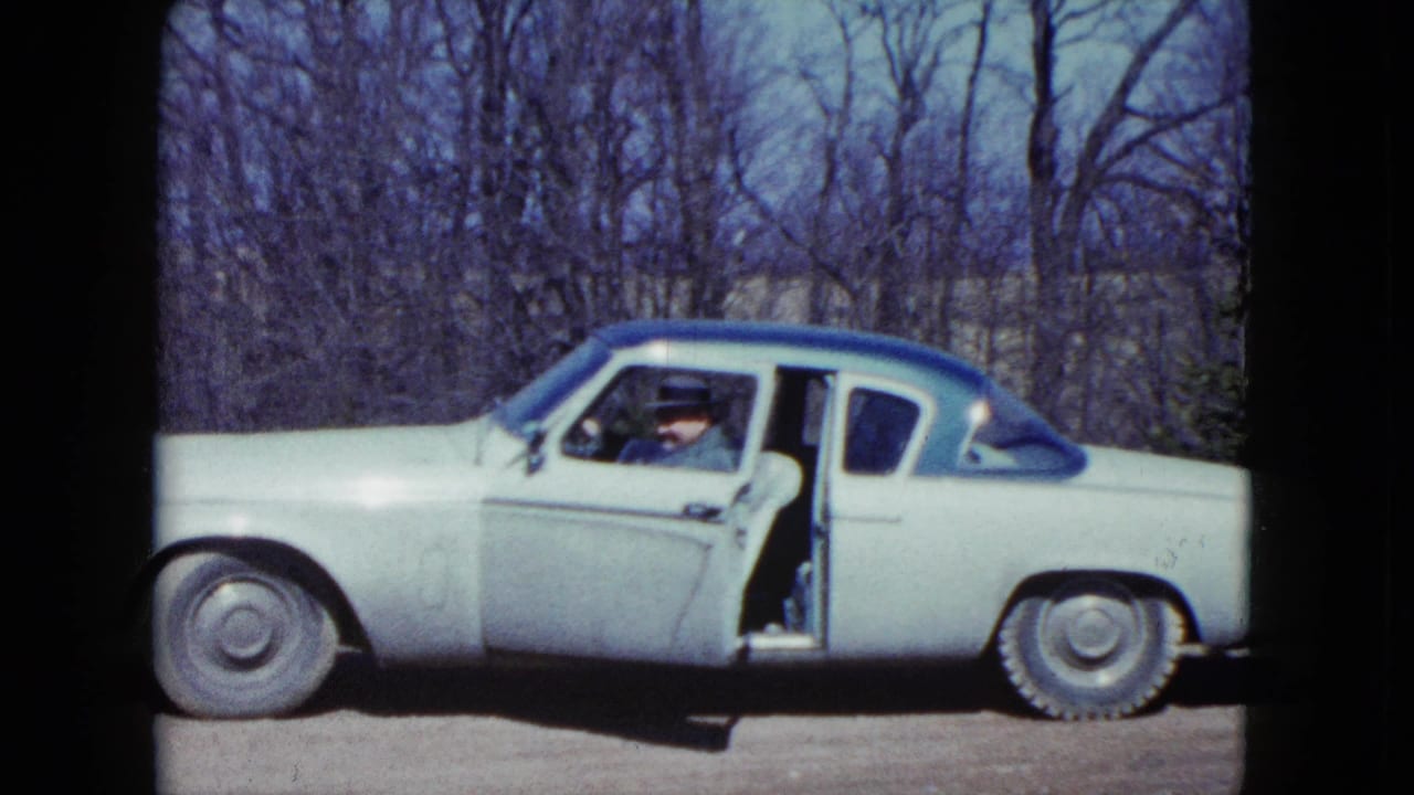 Light-colored classic car with blue roof parked roadside as a man sits by the open door.