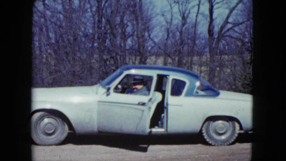 In 1959 Nevada home-movie footage, a suited man appears in a light classic car with a blue roof beside leafless trees.