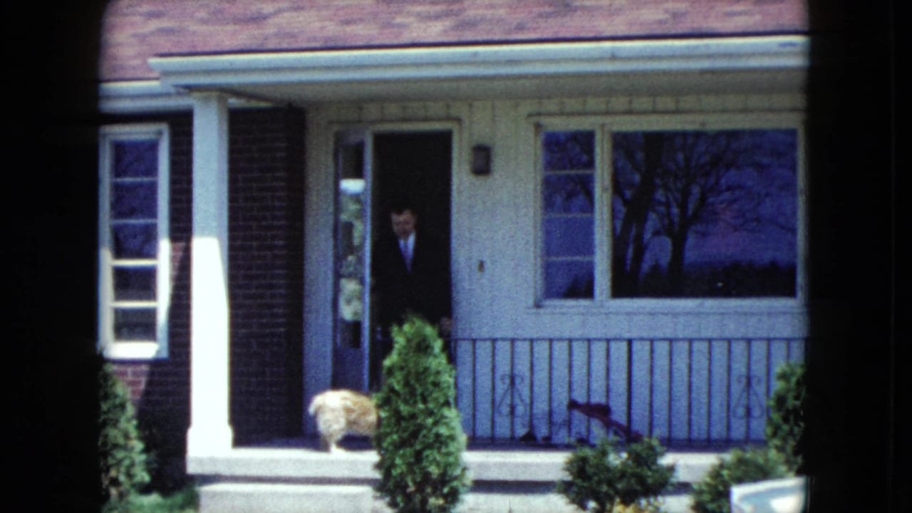 Man in a dark suit stands in the doorway of a suburban house near a small dog on the porch.