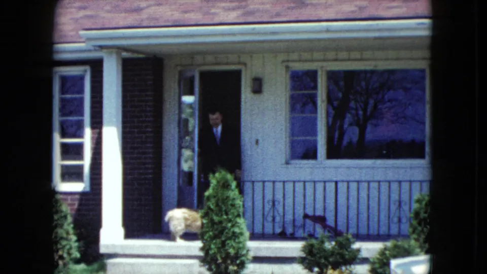 A suited man stands at a suburban front door in the 1959 Nevada clip while a small light-colored dog waits on the porch.