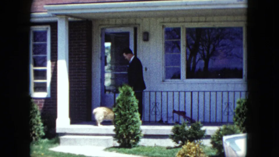 The suited man stands on the suburban porch in Nevada metadata footage, looking down toward a small dog near the entrance.