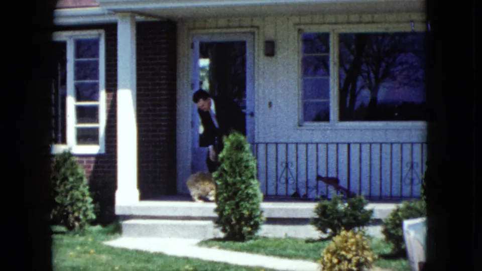 A suited man bends toward the small dog on a suburban porch, framed by shrubs, railing, and a large front window.