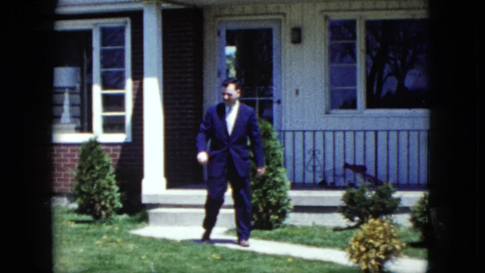 A suited adult walks across the front lawn of a 1959 Nevada suburban house, with porch railings, windows, and shrubs behind him.