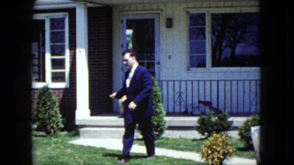 The suited man continues past the front of the Nevada home in 1959, with a porch, railing, windows, shrubs, and lawn visible behind him.