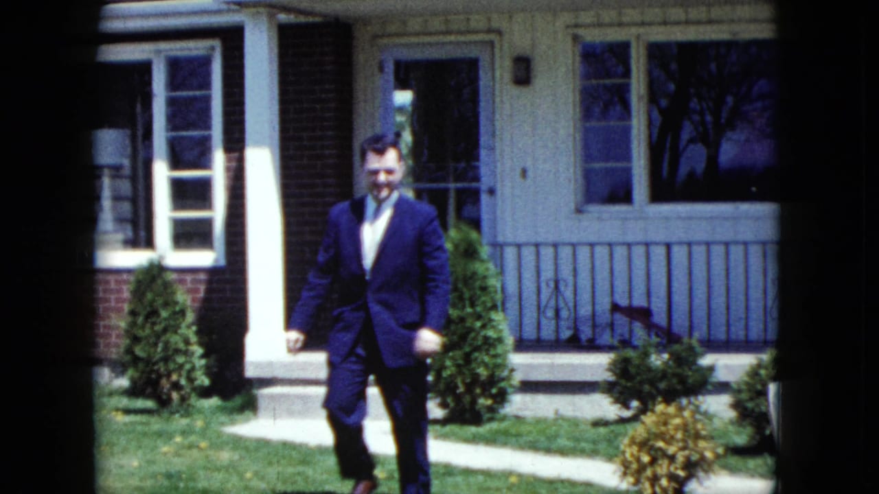 Man in a dark suit walks across a front lawn toward the camera.