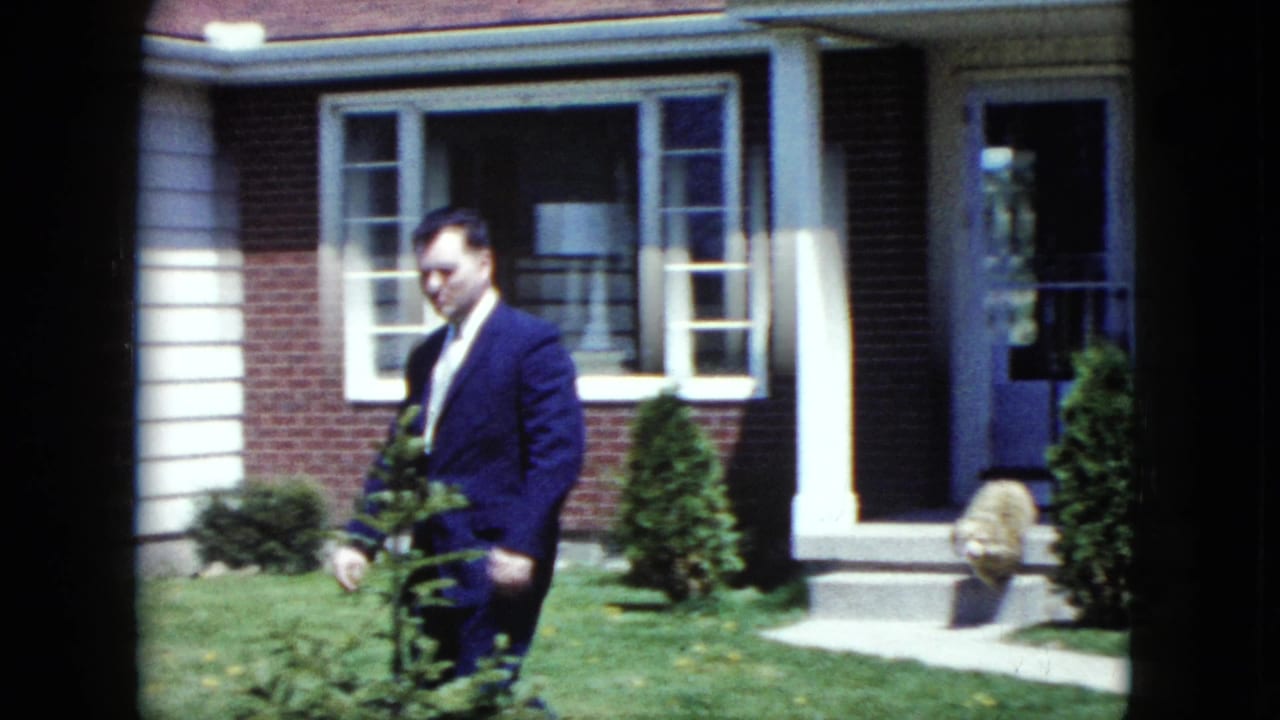 Man in a suit walks past shrubs outside a house with an open doorway.