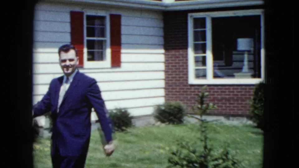 A suited man approaches the camera beside a mid-century suburban house with red shutters and a maintained lawn.
