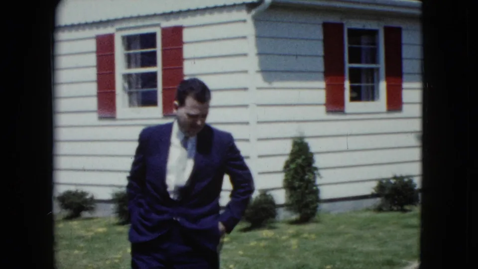 The suited man looks downward while crossing the lawn beside a light-sided house with red shutters and shrubs.