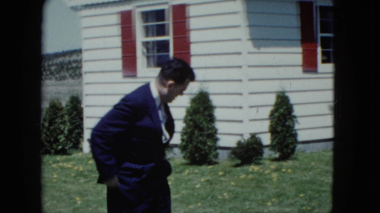 Man in a dark suit bends forward on a lawn beside a white house with red shutters.