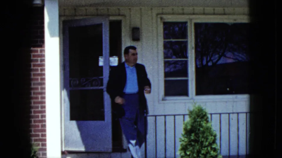 A man appears at the doorway of a Nevada house exterior, wearing a dark jacket and light shirt in 1959 home-movie footage.