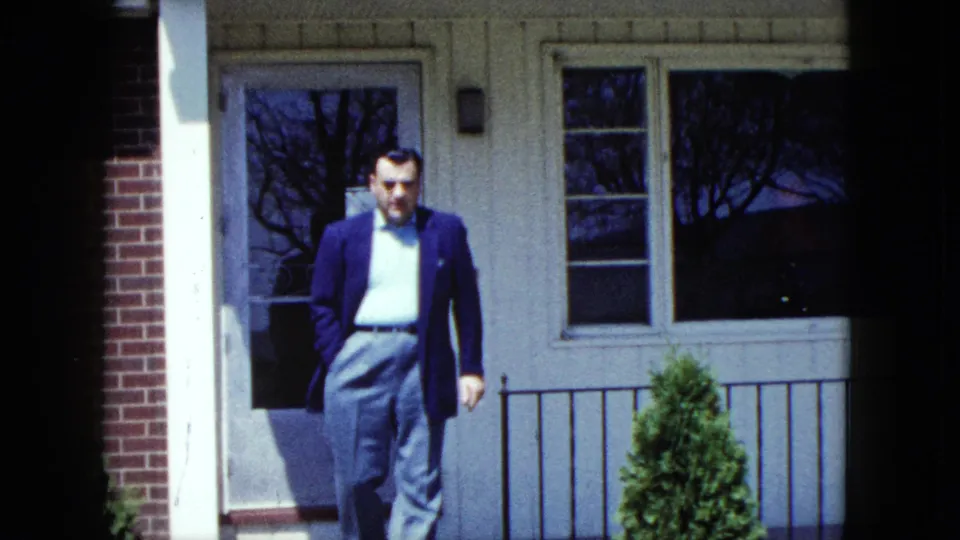 The man walks away from the front entrance of a suburban Nevada house, with windows, railing, and shrubbery behind him.