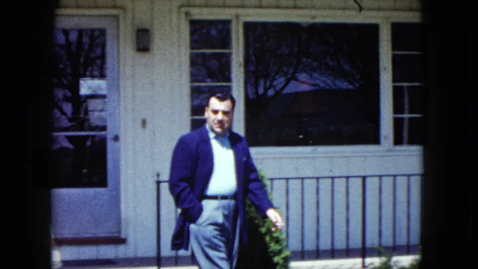 A man in a blue-toned jacket and grey trousers walks past the front of a Nevada house in 1959 home-movie footage.