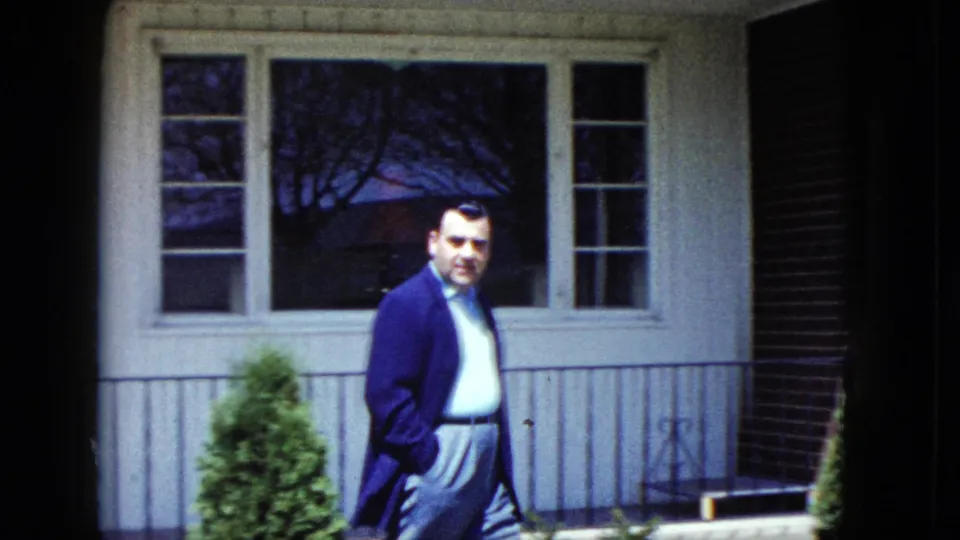 In 1959 Nevada home-movie footage, an adult man in a dark coat walks past a white suburban house exterior.