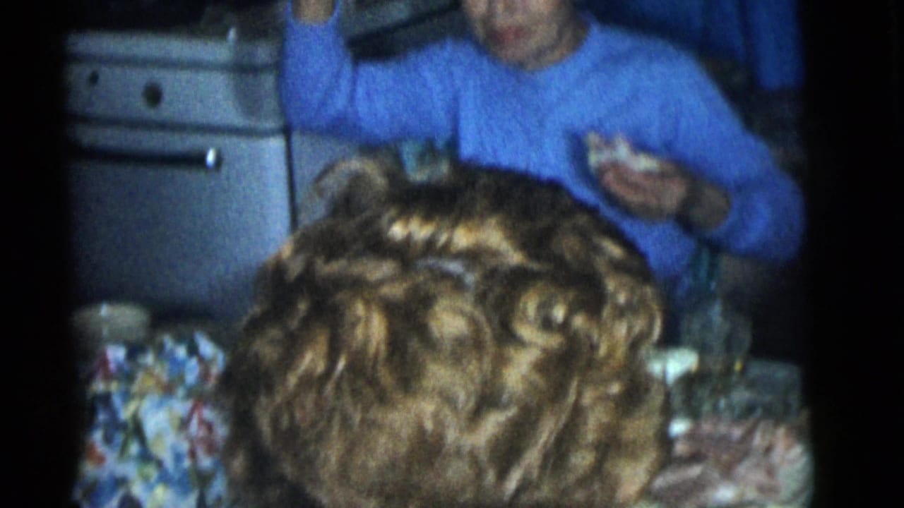 A woman in a blue sweater eats and gestures at a Camden kitchen table, with a white stove and another person partly visible in the foreground.