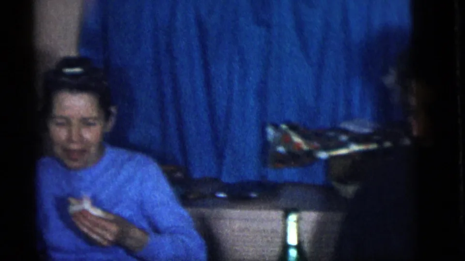 The same woman in a blue sweater sits near blue curtains, holding food during the 1960 Camden home lunch gathering.