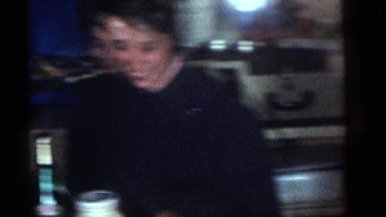 Blurred adult seated indoors near a table, with dark clothing and household fixtures behind.