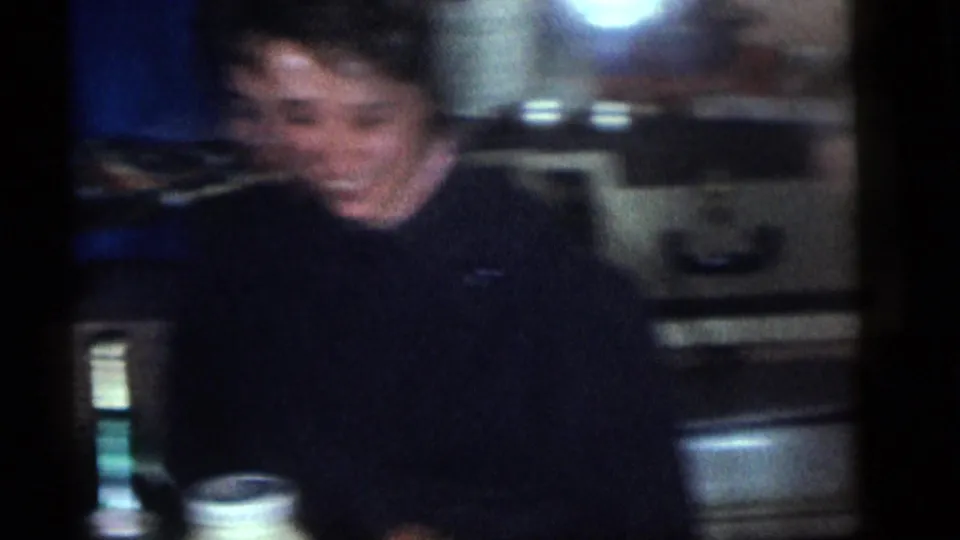 Soft, blurred 1960 Camden home-movie frame of an adult seated indoors near a table, with a domestic background and dim lighting.