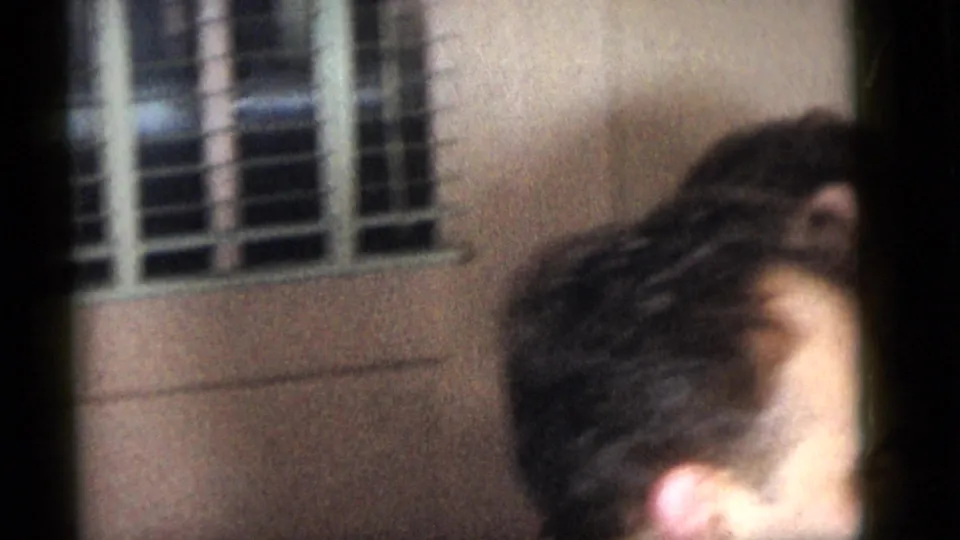 Partial close-up from the Camden 1960 home movie showing an adult at the edge of frame near window blinds inside a private room.