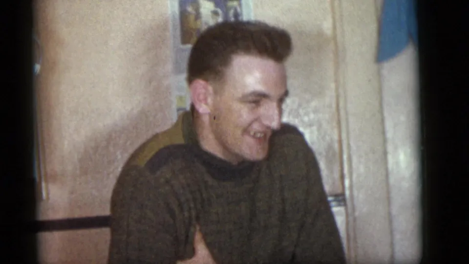 A smiling adult man in a dark knit sweater sits indoors in the 1960 Camden home-movie gathering, framed in soft 8mm light.