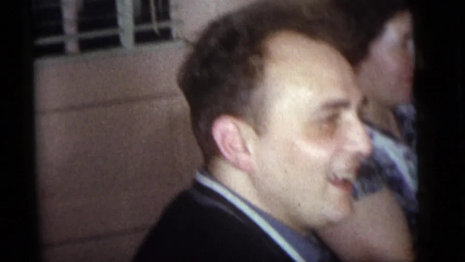 Profile view of an adult man laughing or smiling beside another person in the Camden 1960 home-movie interior gathering.