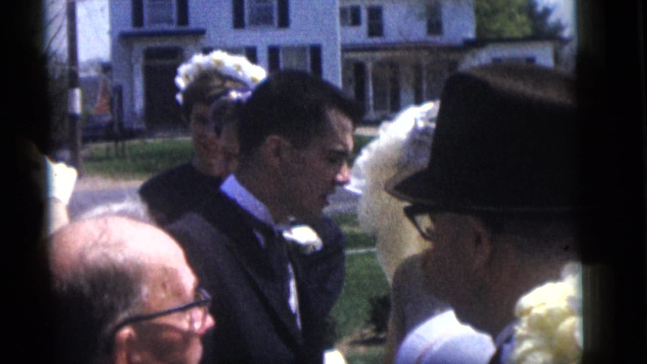 Groom and guests stand outdoors near a bride in a white veil.