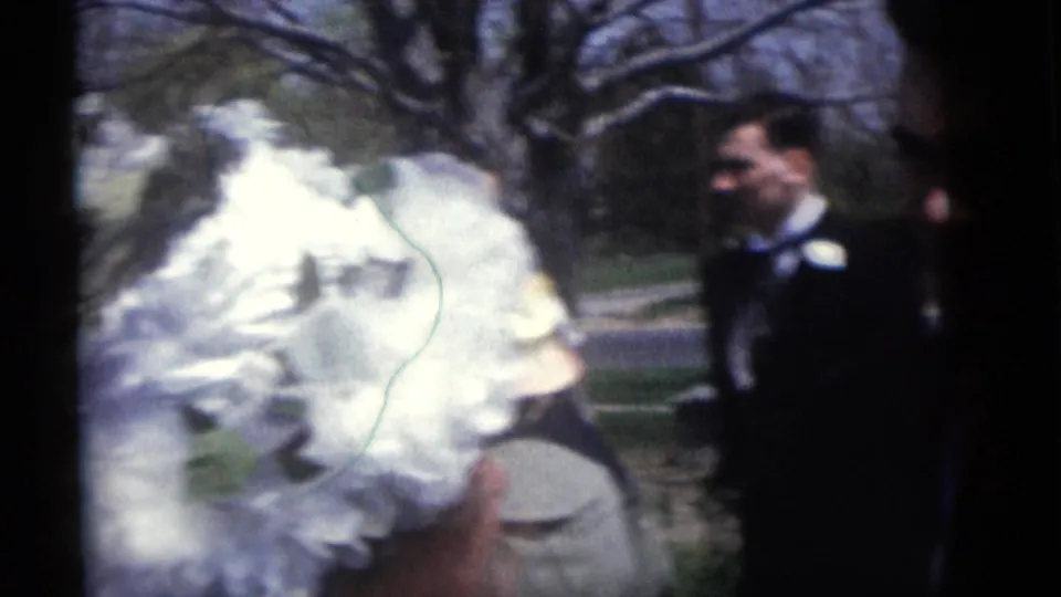 A white bridal garment fills the foreground as a suited man stands in the grassy outdoor setting of the Camden wedding footage.