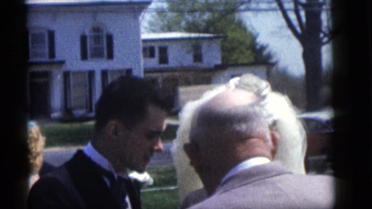 A groom, bride, and older male figure are seen during the Camden outdoor wedding ceremony, with a white house behind them.