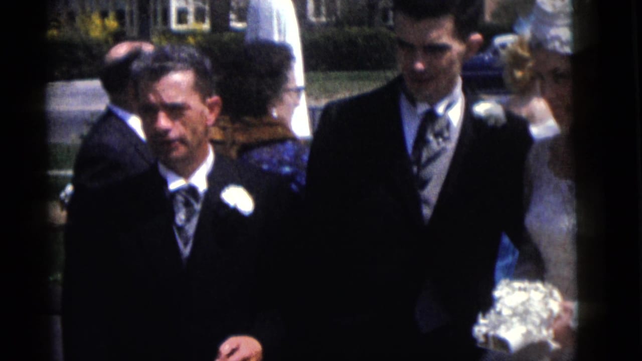 1960 Camden home-movie frame showing suited men with boutonnieres and wedding guests near a bride on a sunny residential lawn.