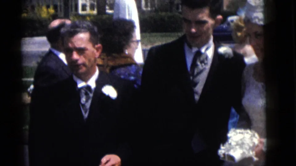 1960 Camden home-movie frame showing suited men with boutonnieres and wedding guests near a bride on a sunny residential lawn.