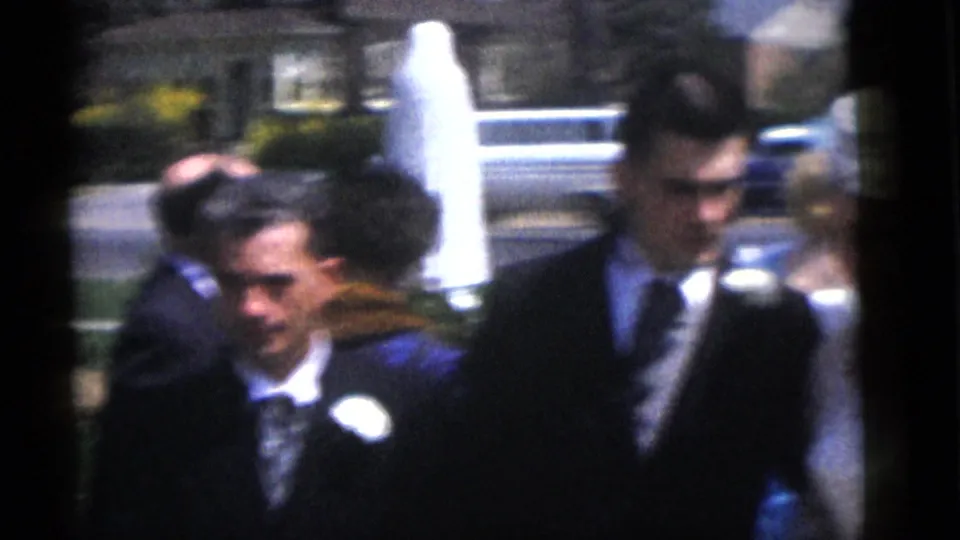1960 Camden wedding party members and guests pass through an outdoor residential setting, framed by houses and a street.