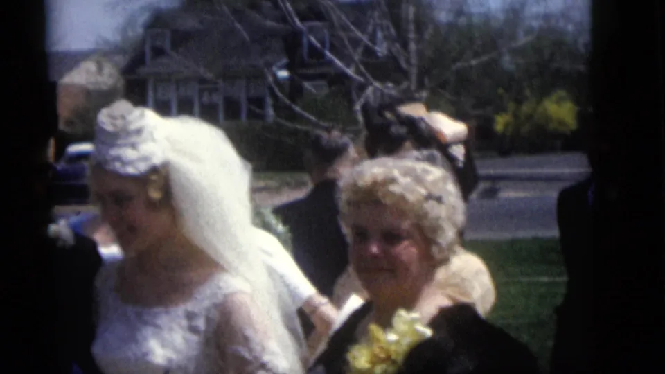 Bride in a white lace gown and veil walks beside a woman with flowers during the 1960 Camden outdoor family wedding scene.