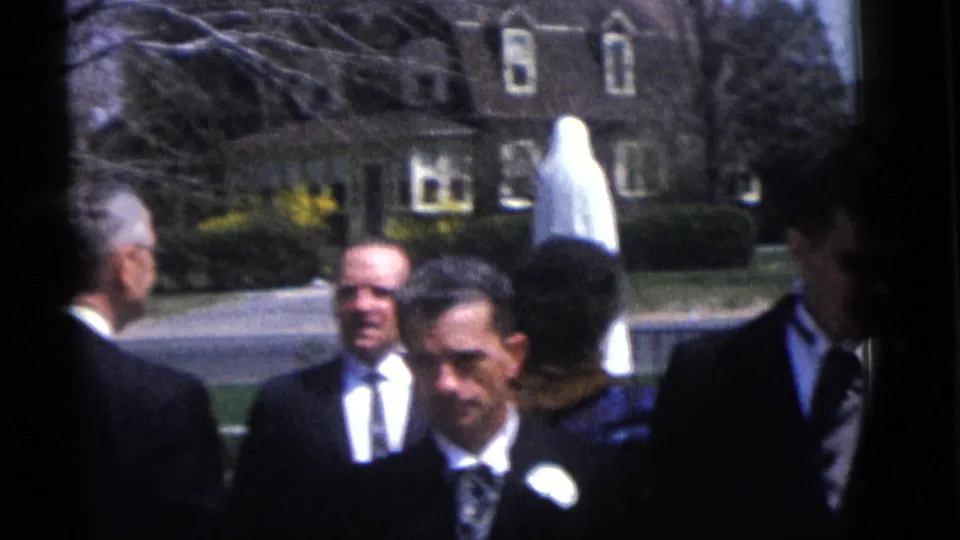 1960 Camden home-movie frame of suited wedding guests gathered on a lawn, with residential houses and a street behind them.