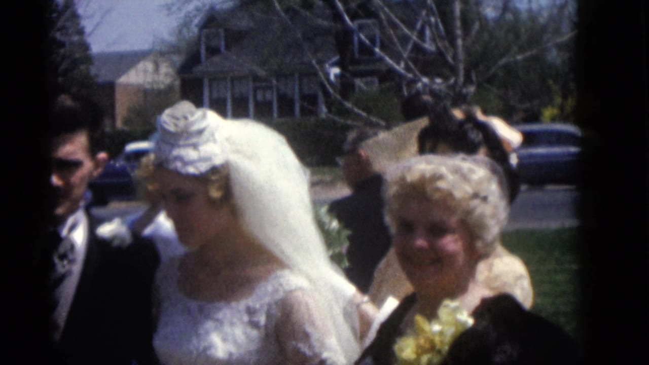 Bride in veil walks beside groom and smiling guest outside a house.