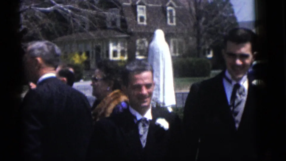 1960 Camden wedding home-movie frame showing suited men, guests, a veiled bride partly visible, and houses across a residential street.
