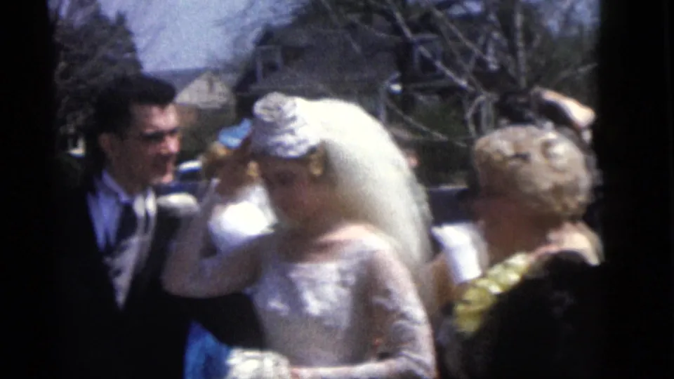 1960 Camden wedding frame with the bride in a lace gown and long veil adjusting her headpiece beside the groom outdoors.
